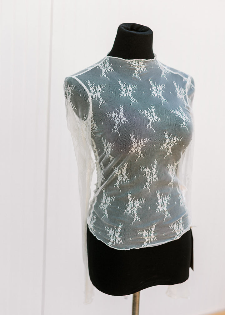 Mannequin wearing a sheer, long sleeve lace top with floral, mock neckline and slight ruffle detail.