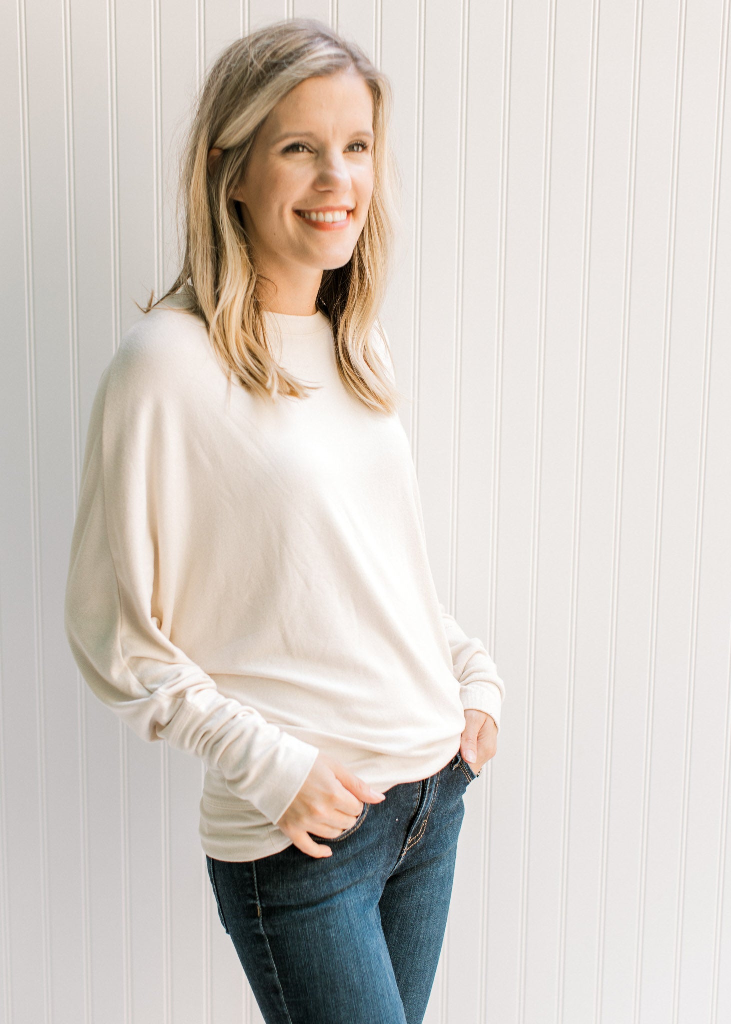 Model wearing jeans with a cream sweater with batwing sleeves, round neck and polyester material. 
