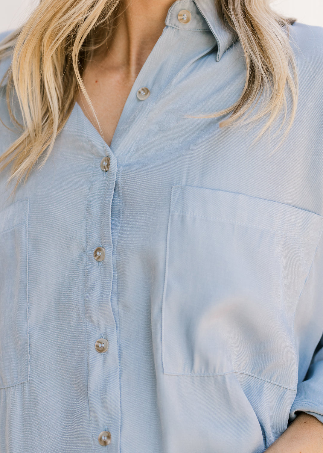 Close up of button front and patch pockets on a light blue top with a traditional collar. 
