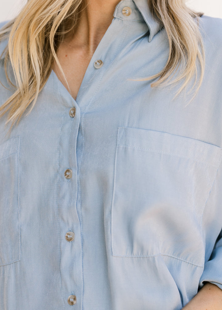 Close up of button front and patch pockets on a light blue top with a traditional collar. 
