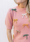 Model wearing a dusty pink sweater with cream, taupe and green bows and short sleeves. 