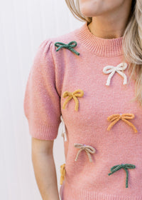Model wearing a dusty pink sweater with cream, taupe and green bows and short sleeves. 