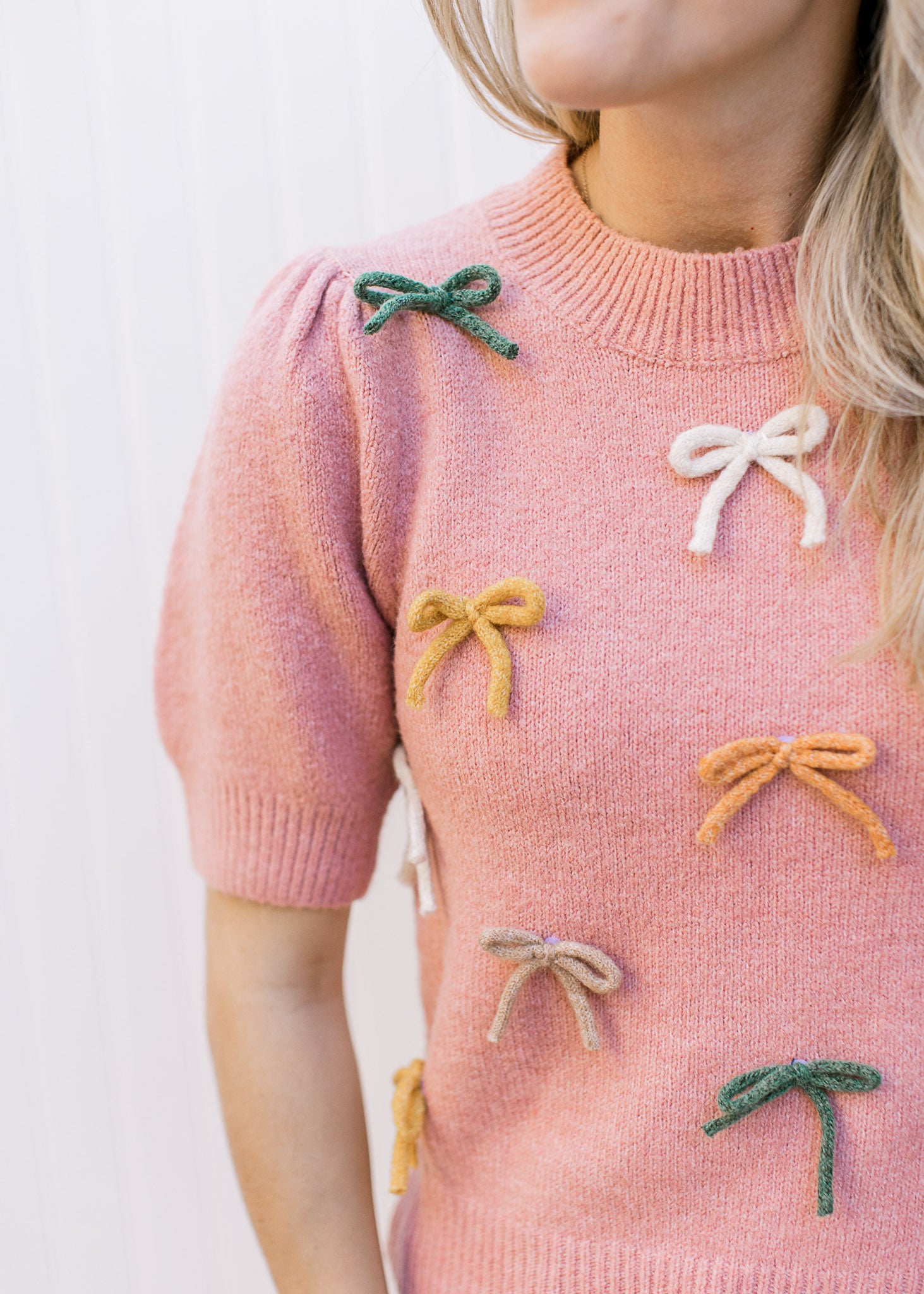 Model wearing a dusty pink sweater with cream, taupe and green bows and short sleeves. 