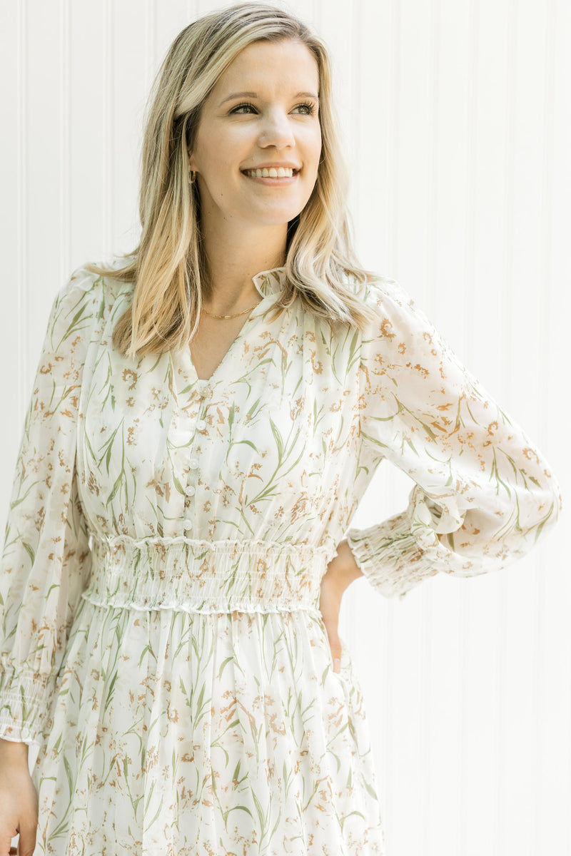 Model wearing a cream dress with a tan floral pattern, elastic waistband and sheer long sleeves. 