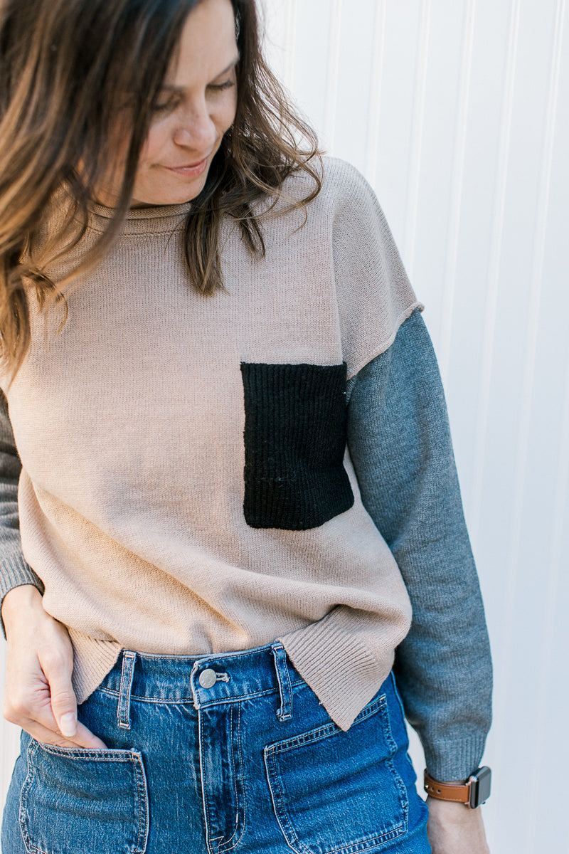 Close up of black patch pocket on a camel colored sweater with gray sleeves. 