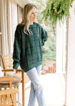 Model wearing a hunter green sweater with black window pane pattern and long sleeves. 