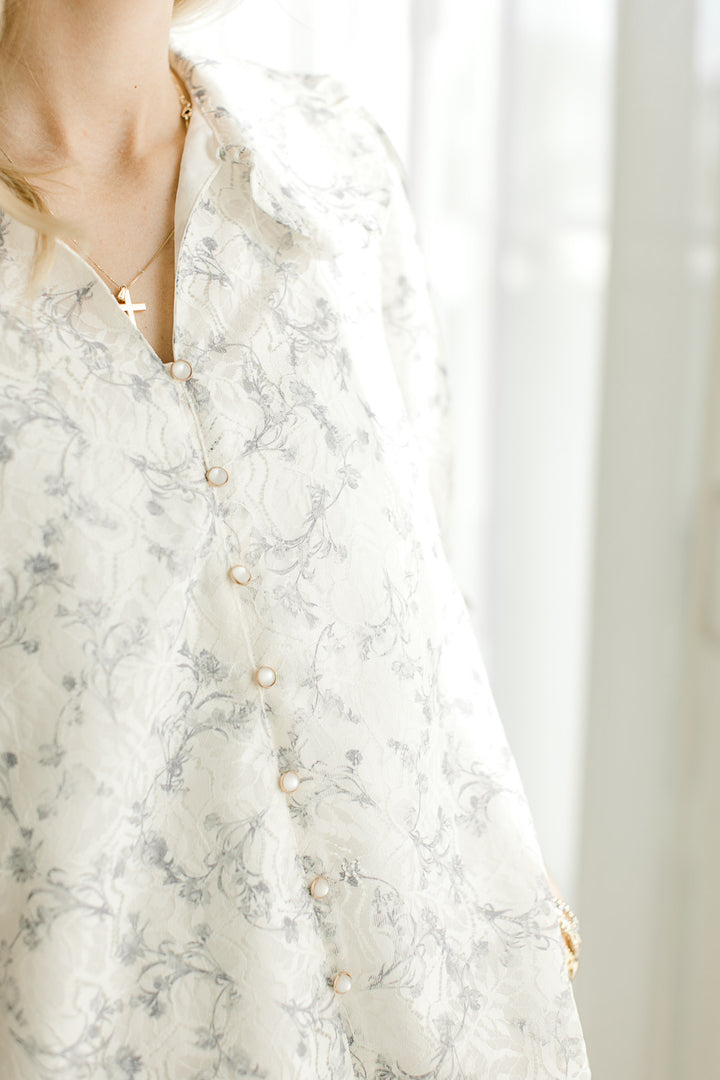 Close up of v-neck and pearl buttons and an off-white top with gray floral and lace overlay. 
