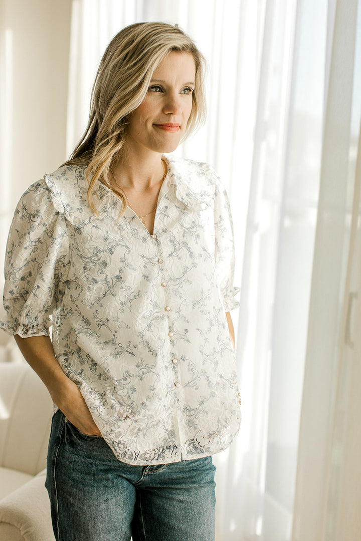 Model wearing a short sleeve off-white top with gray floral, lace overlay, flap collar and buttons.