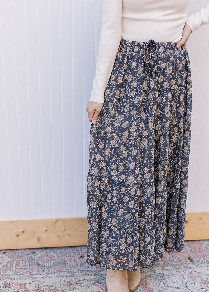 Model wearing a flowy, slate blue midi with tan floral, elastic waist with tie, tiers and pockets. 