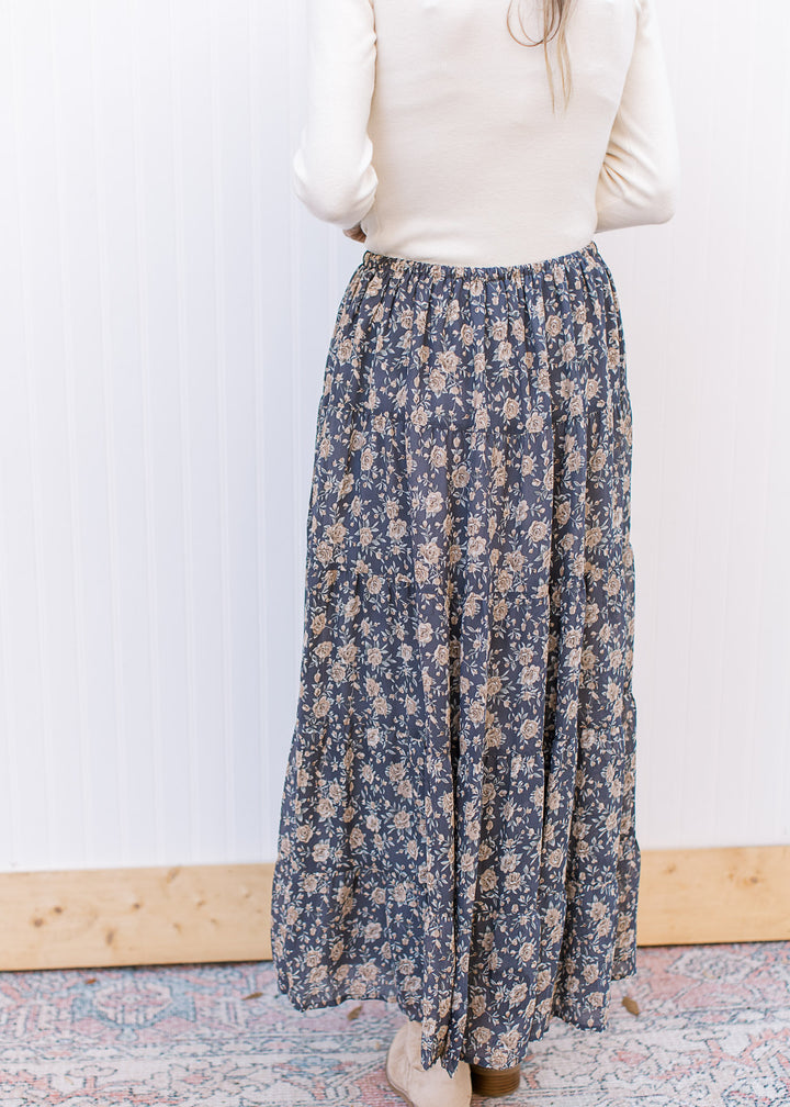 Back view of Model wearing a flowy, slate blue midi with tan floral, elastic waist and pockets. 