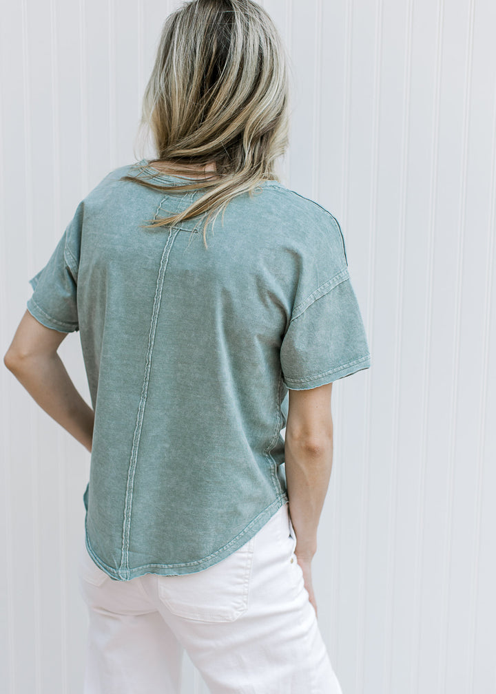 Back view of Model wearing a slate blue top with a v-neck, short sleeves and a raw hemline.