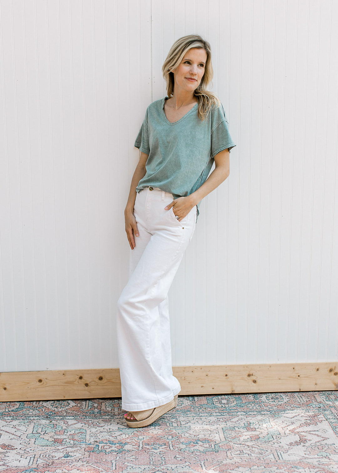 Model wearing white pants and a slate blue top with a v- neck, short sleeves and a raw hemline.