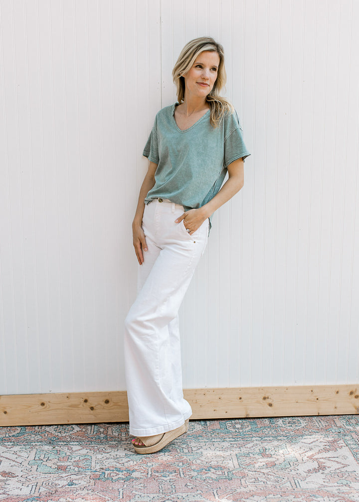 Model wearing white pants and a slate blue top with a v- neck, short sleeves and a raw hemline.