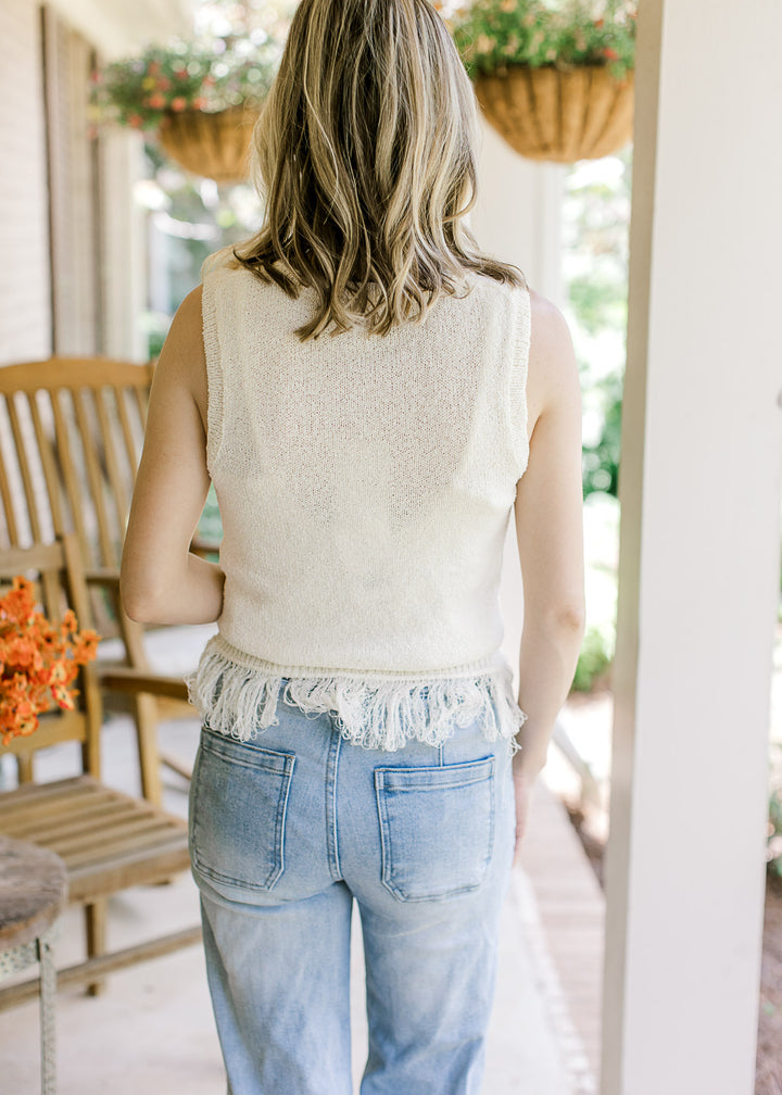 Back view of Model wearing a cream sleeveless blouse with a round neck and fringe hem detail.