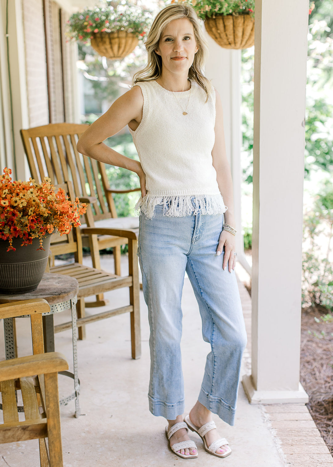 Model wearing jeans, sandals and a cream sleeveless blouse with a round neck and fringe hem detail. 