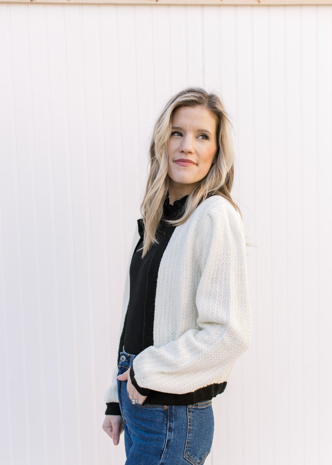 Side view of Model wearing a cream sweater with a black cuff, hem and neck and an open front.