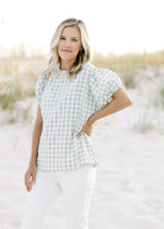 Model wearing a sage and white short sleeve plaid top with smocked detail at the shoulder and neck. 