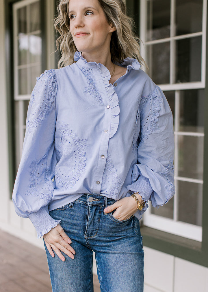 Model wearing a long sleeve, soft blue top with floral medallions, button front and ruffle detail. 
