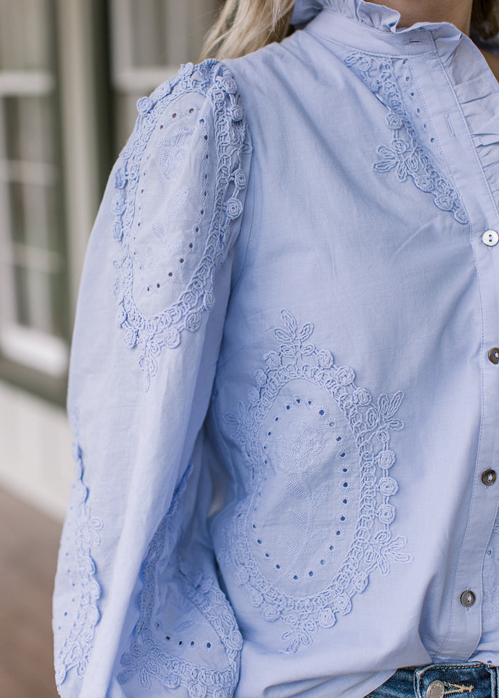 Close up of tone-on-tone floral medallions and ruffle detail at neck of a soft blue long sleeve top.