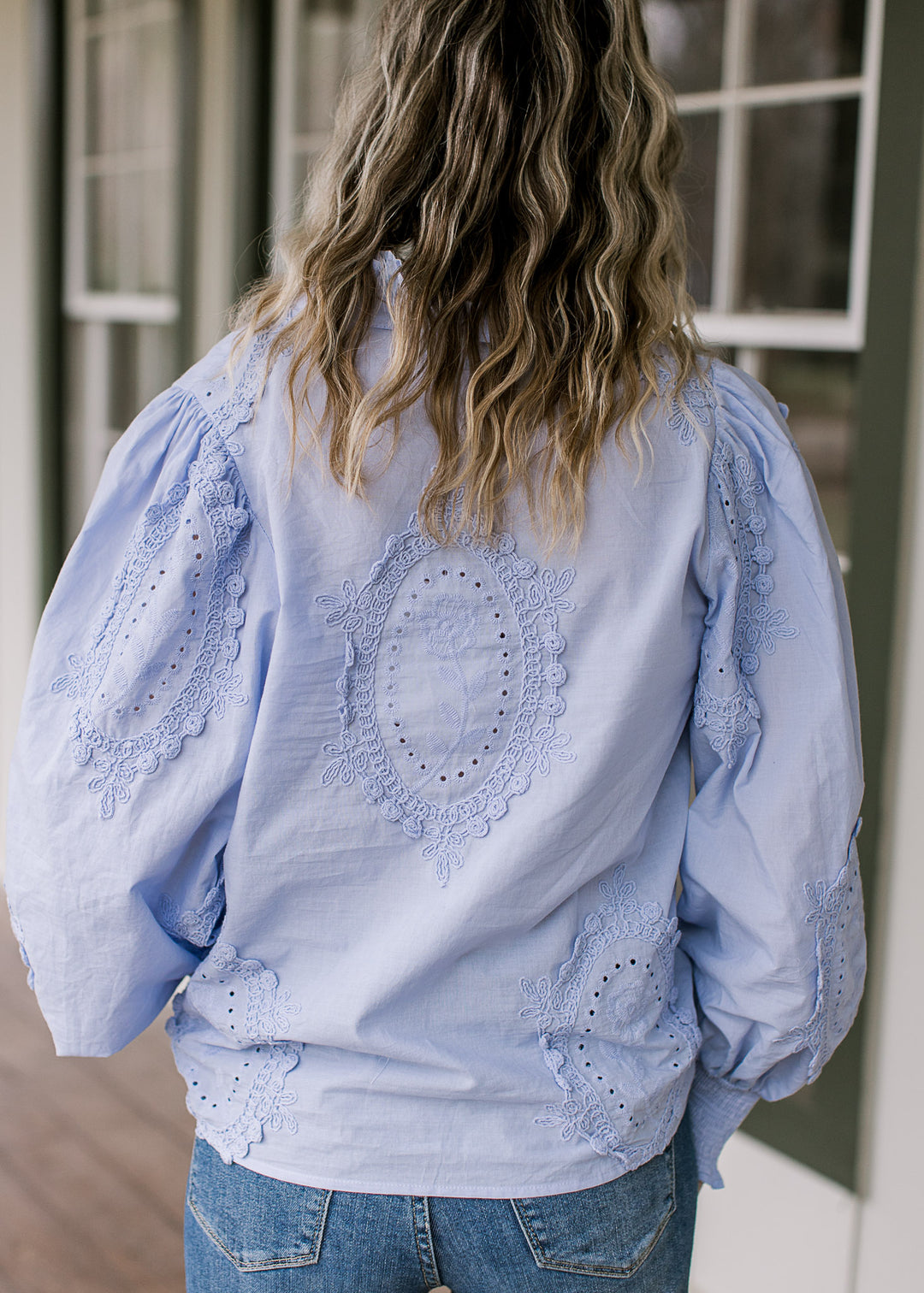 Back view of Model wearing a soft blue top with tone-on-tone floral medallions and long sleeves. 