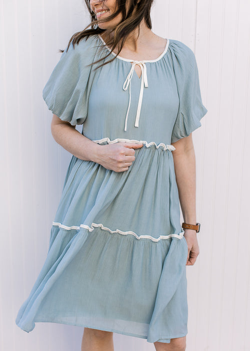 Model wearing a pale blue-green knee length dress with cream piping at neckline and each tier.