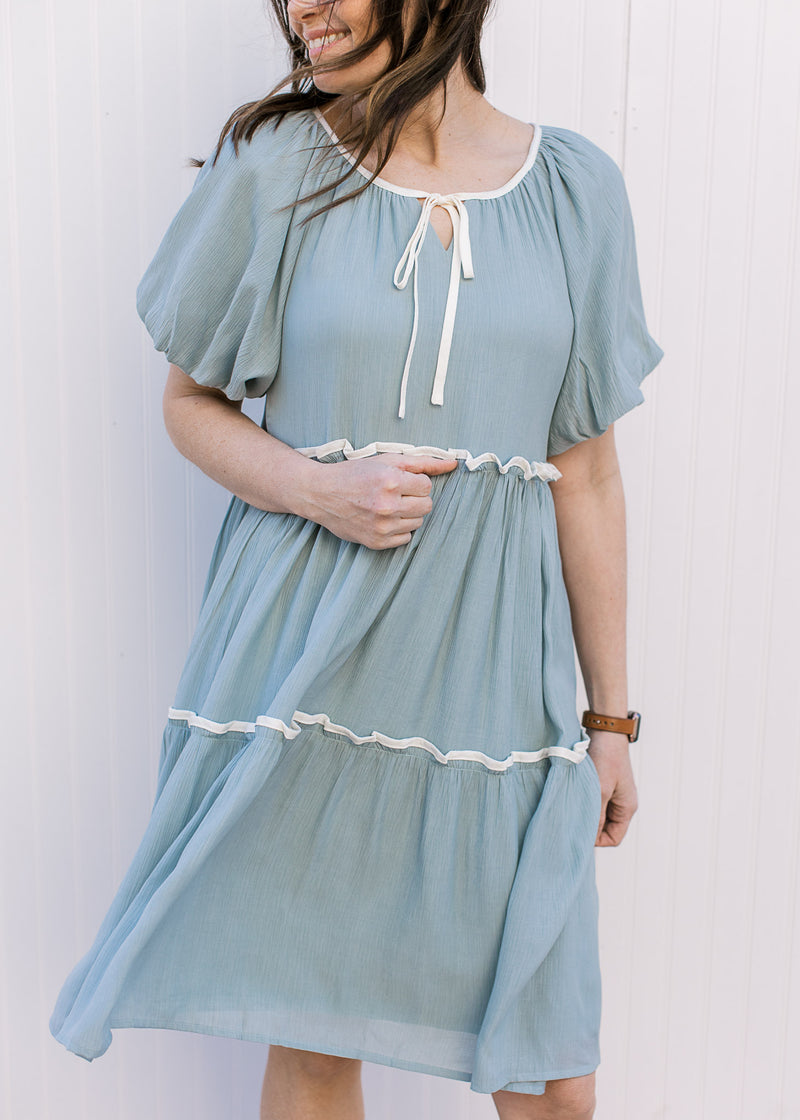 Model wearing a pale blue-green knee length dress with cream piping at neckline and each tier.