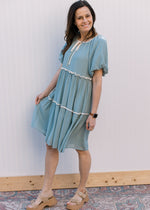 Model wearing sandals and a pale blue-green dress with cream piping and tie keyhole at neck.