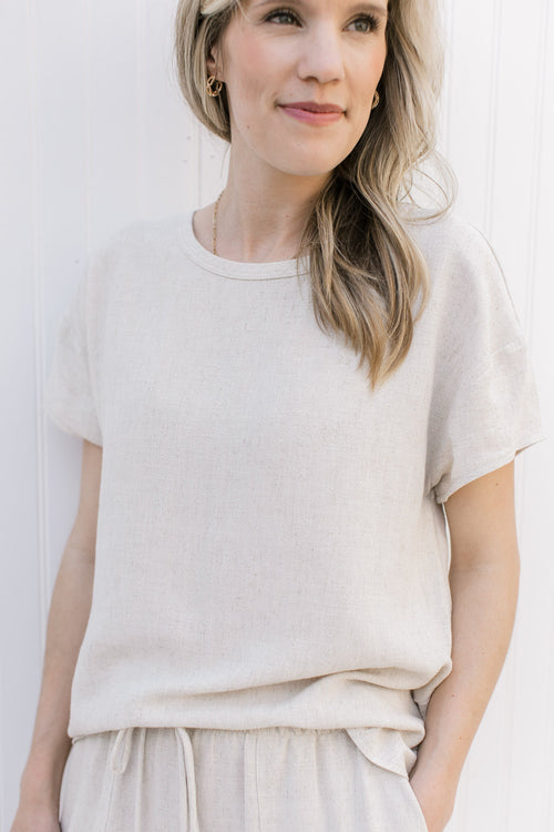 Model wearing a oatmeal colored short sleeve top with a round neck and linen/ rayon blend. 