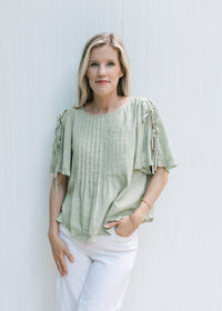 Model wearing a soft green top with ruffle at cuff and hem and tie detail on the short sleeves.