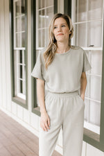 Model wearing a sage jumpsuit with an elastic waist, short sleeves and pockets. 