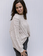 Side view of Model wearing an ivory long sleeve, cable knit sweater with a soft polyester material.