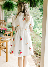 Back view of model wearing a white dress with embroidered flowers, elastic waist and short sleeves.