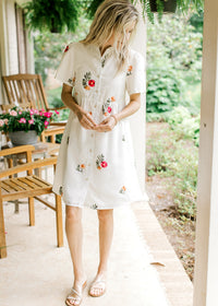 Model wearing a short sleeve button up dress with bright embroidered flowers and a collar.