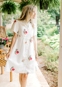 Model wearing a white button up top with embroidered flowers, short sleeves and a collar. 