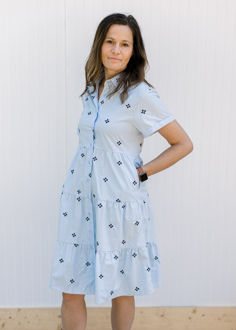 Model wearing a light blue dress with navy embroidered flowers, button front and short sleeves.