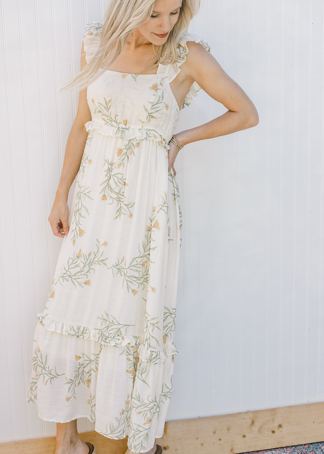 Model wearing a sleeveless, yellowy cream midi with floral, ruffle tiers and a square neck. 