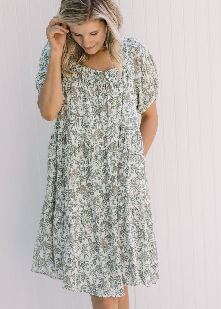 Model wearing a cream above the knees dress with a green floral pattern and pockets. 
