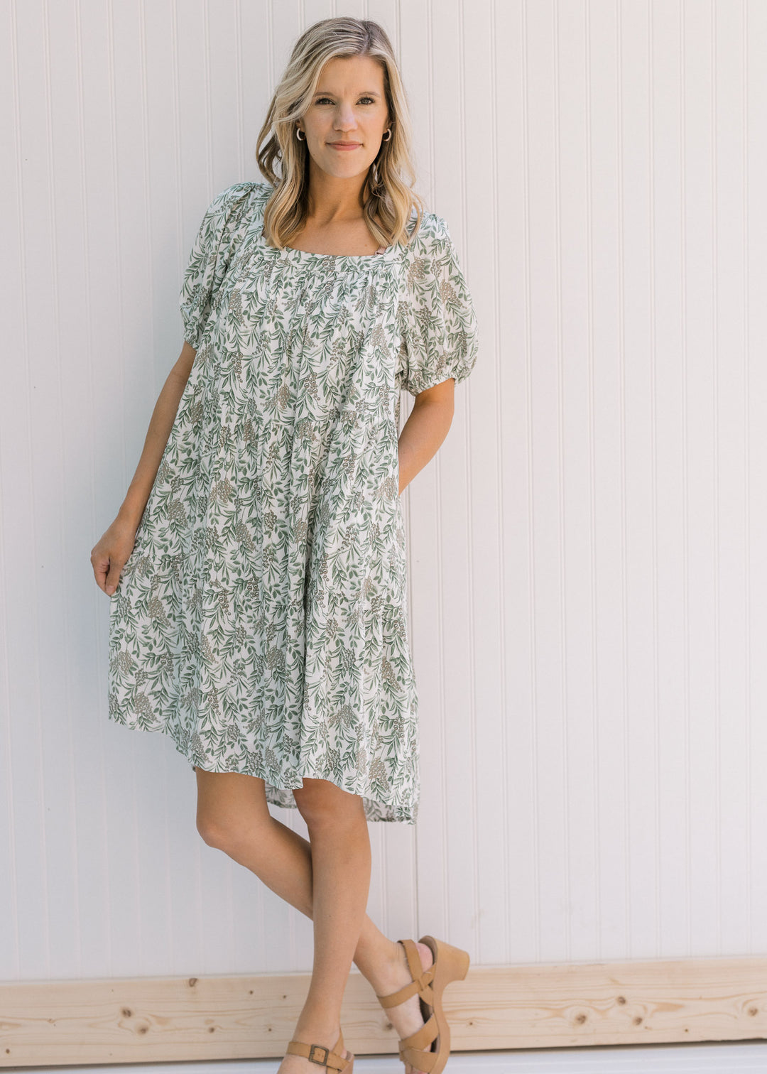 Model wearing a cream above the knee dress with a green floral pattern, a square neck and heels. 