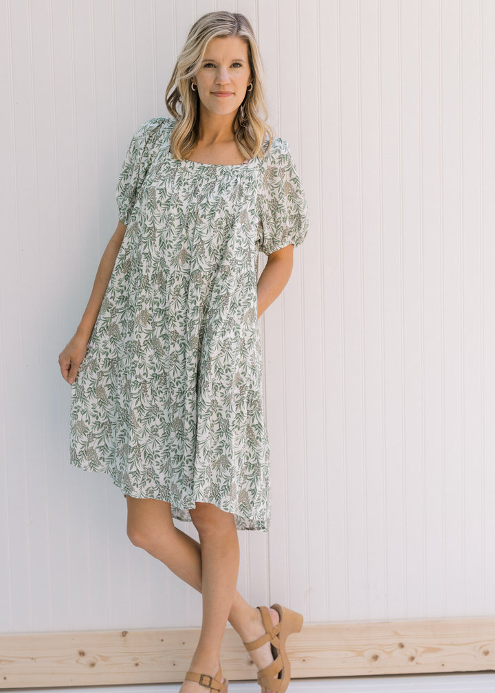 Model wearing a cream above the knee dress with a green floral pattern, a square neck and heels. 