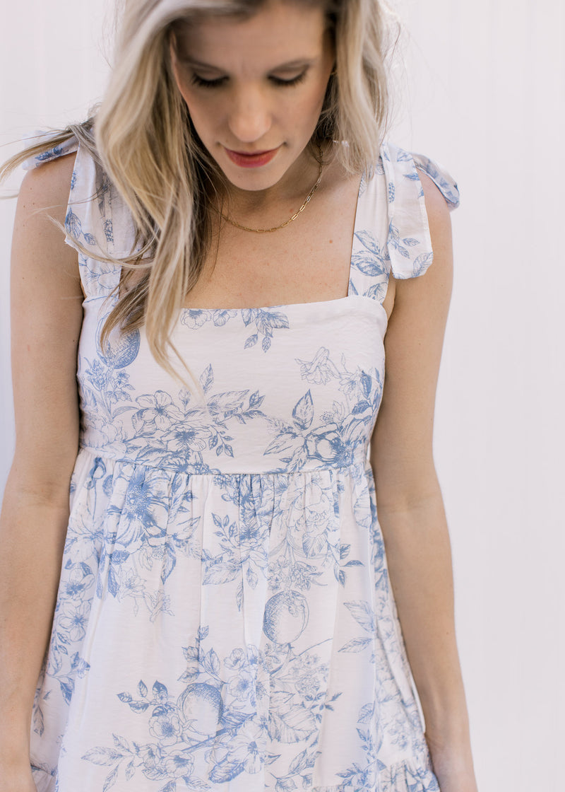 Model wearing a white dress with baby blue floral pattern, square neckline and tie straps. 