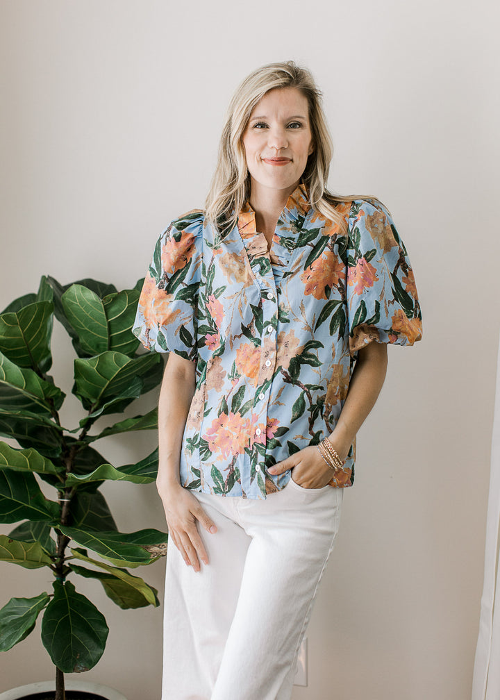 Model wearing white pants and a pale blue top with coral and tan floral and puff short sleeves. 