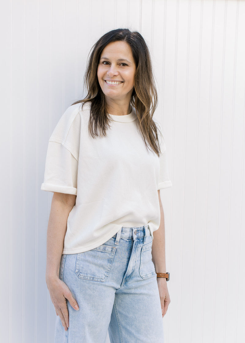 Model wearing a cream ribbed top with cuffed short sleeves and a round neck.