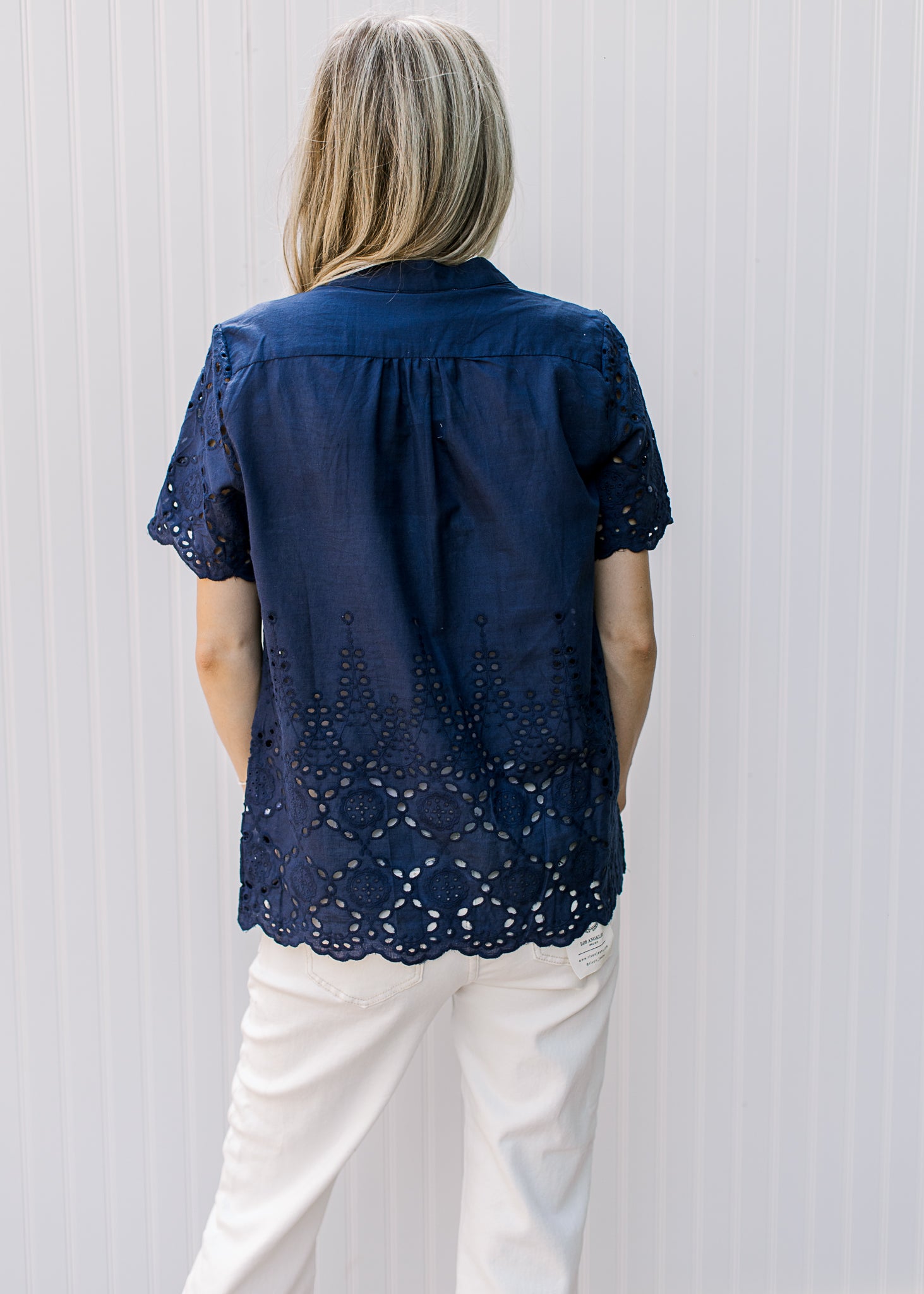 Back of Model wearing a navy button up top with eyelet detail and scalloped short sleeves and hem.
