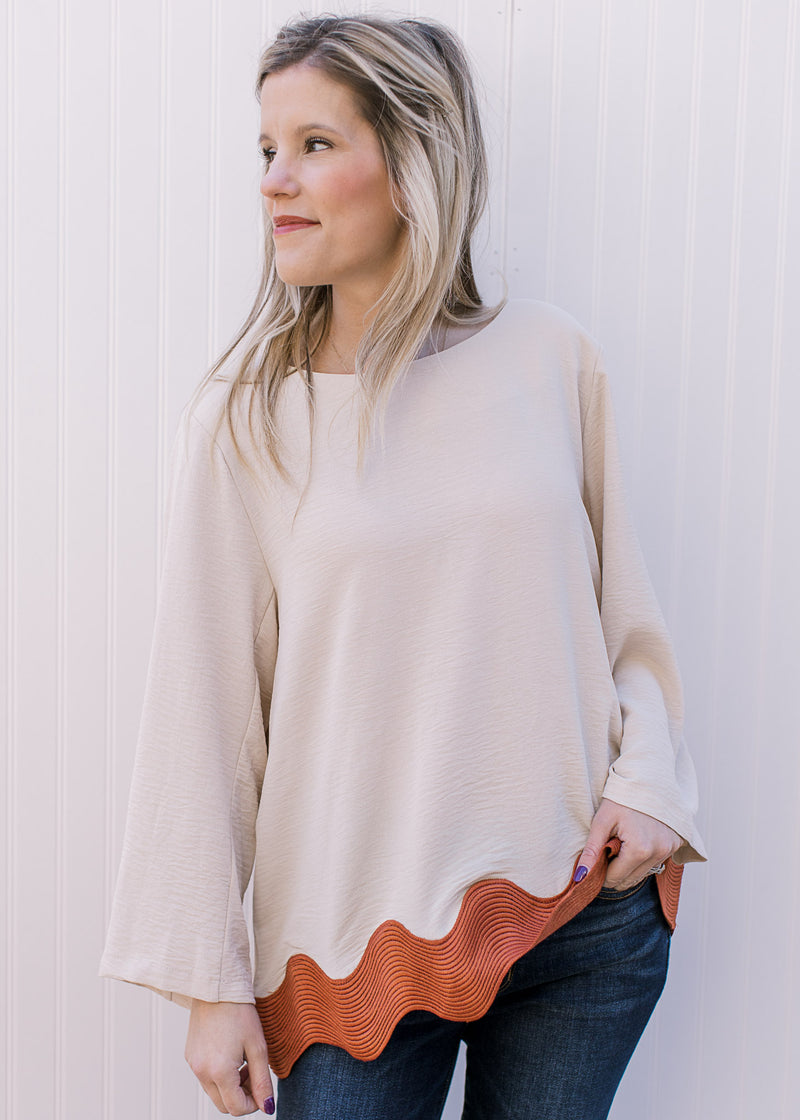 Model wearing a cream top with rust ric-rac hem, 3/4 sleeves, keyhole closure and a round neck.