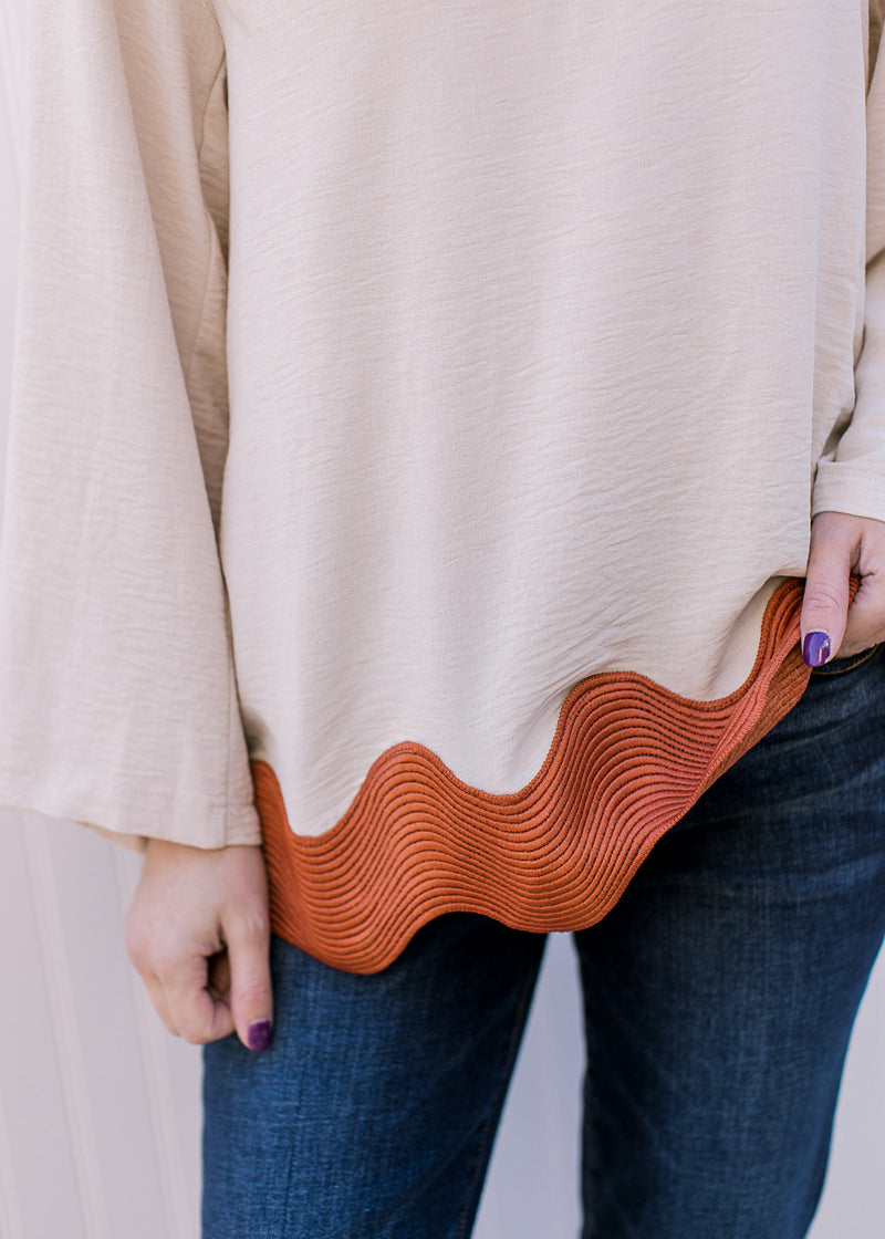 Close up of rust ric-rac hem on a cream top with 3/4 sleeves, keyhole closure and a round neck.