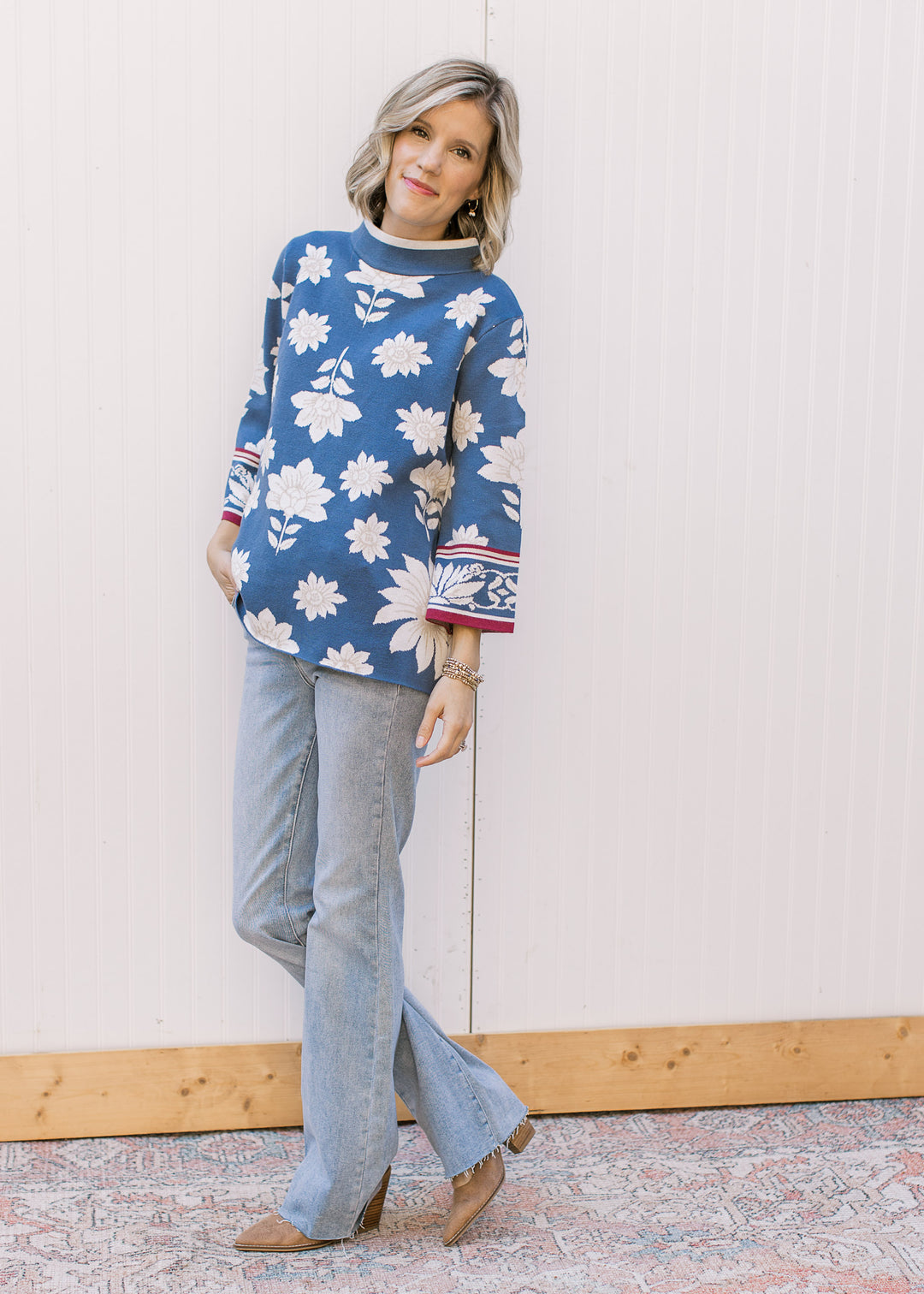 Model wearing jeans, boots and a steel blue sweater with cream and tan floral and mock neck.