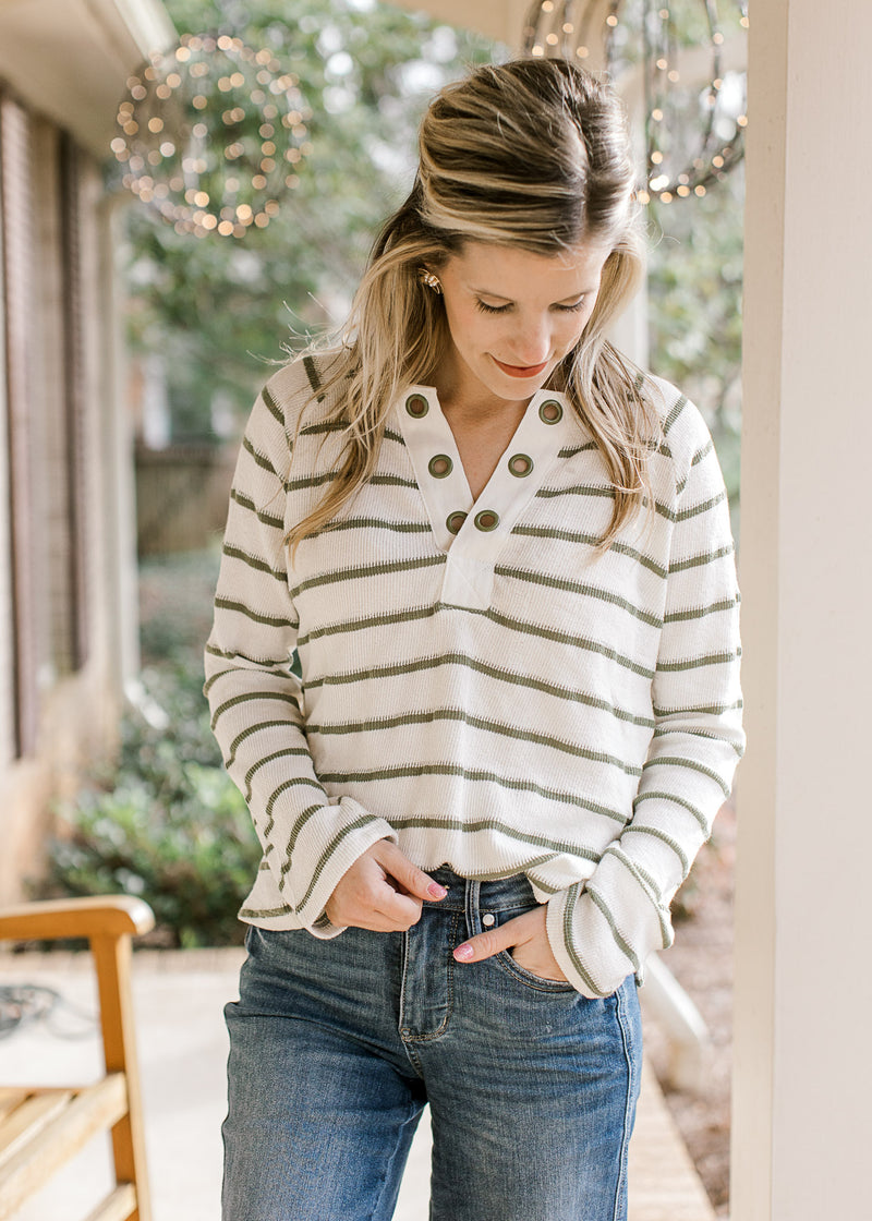 Model wearing a cream top with olive stripes, long sleeves and eyelets along the v-neck. 