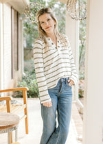 Model wearing jeans and a cream top with olive stripes, long sleeves and eyelets along the v-neck. 