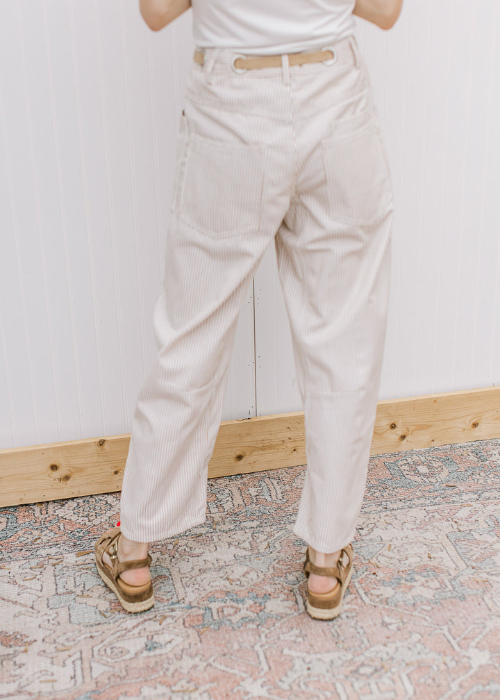 Back view of a model wearing cream barrel leg pants with tan pinstripes and a woven belt. 