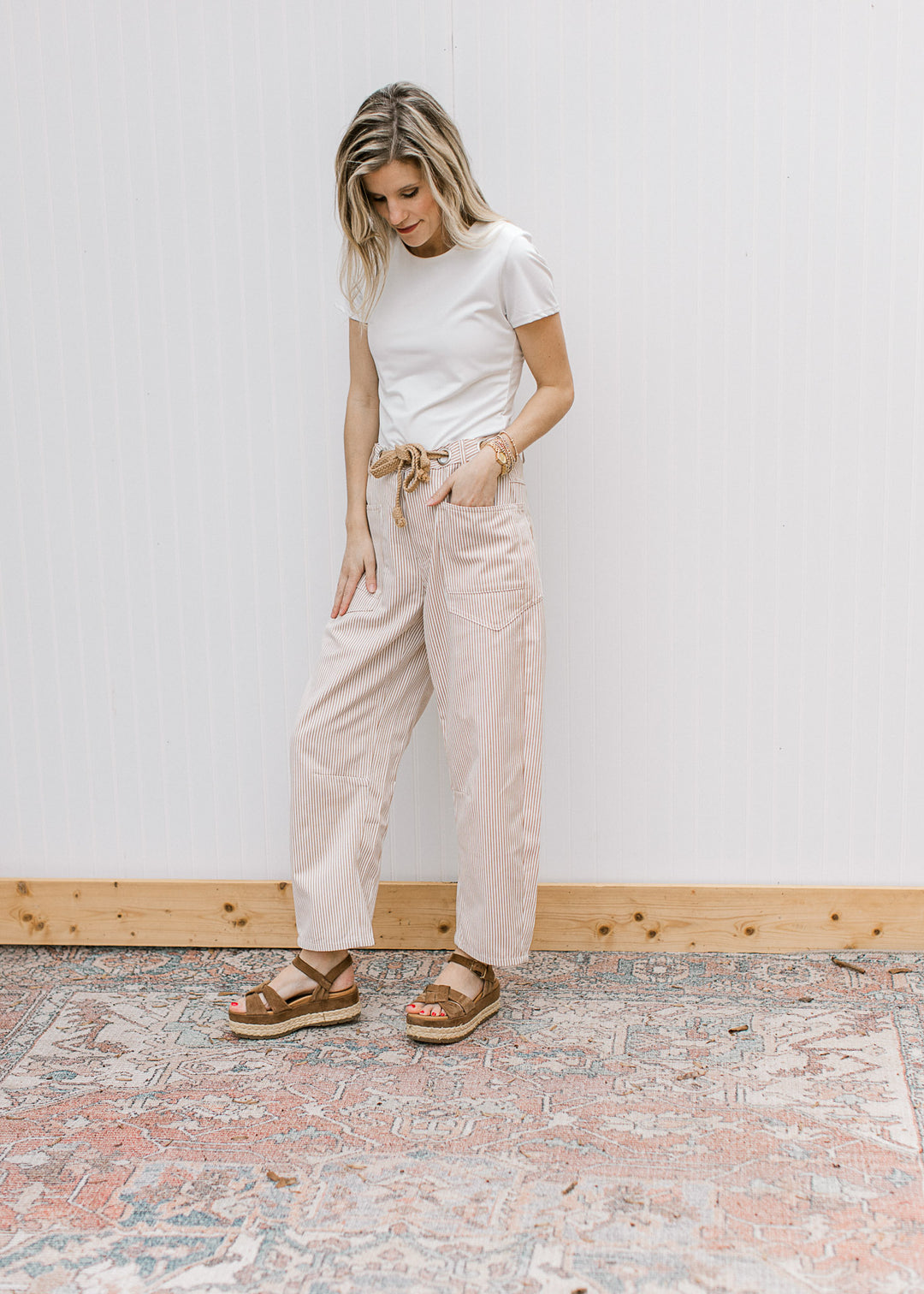 Model wearing a white top, sandals and cream barrel leg pants with tan pinstripes and a belt.